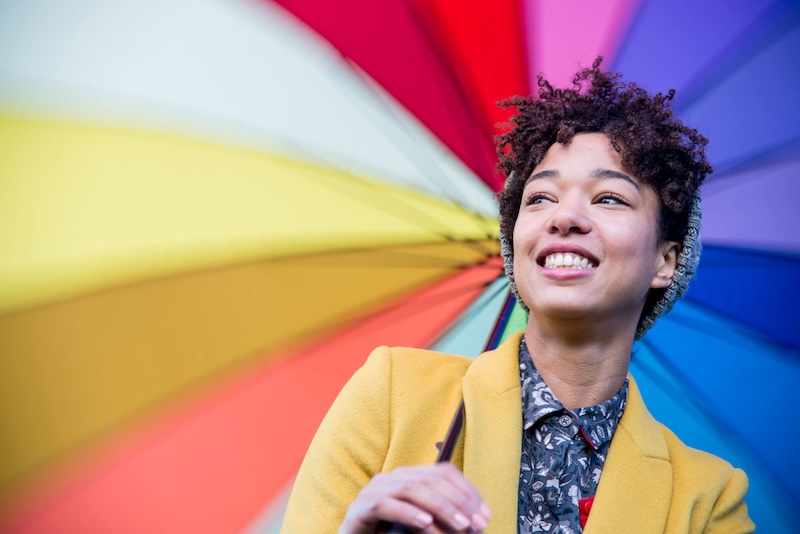 A young lady is standing under a multi coloured umbrella